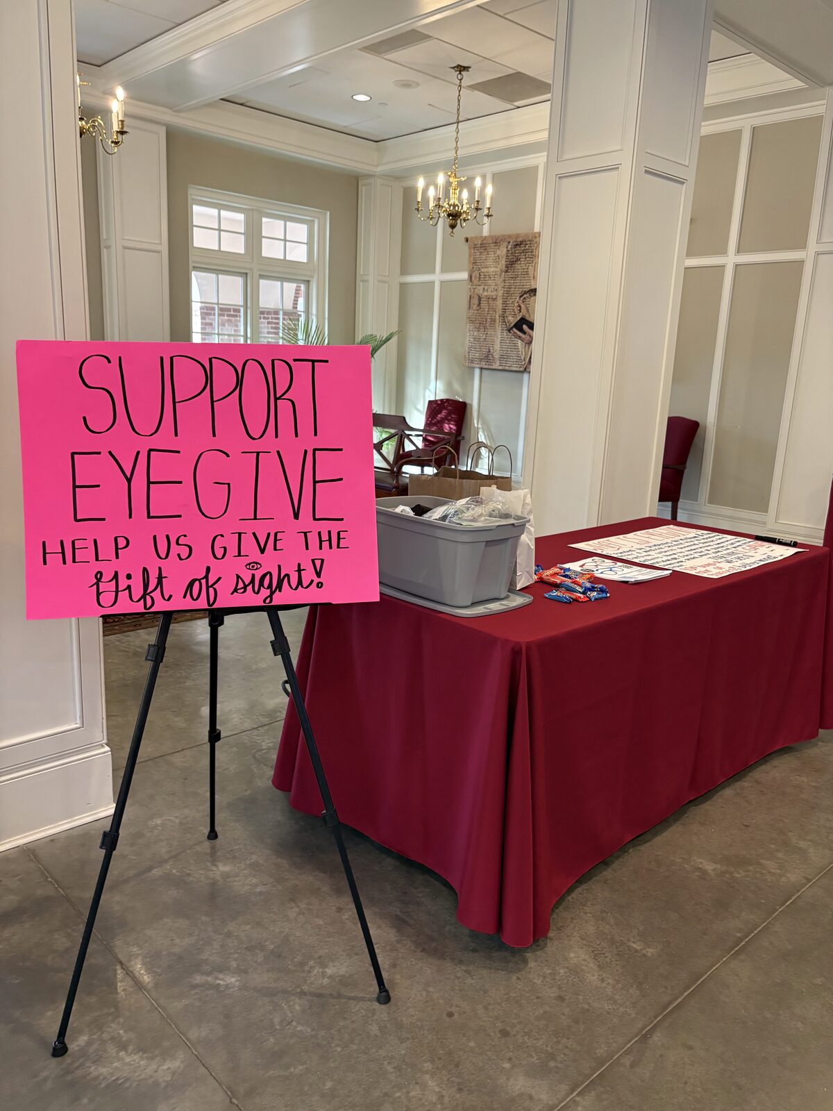 An EyeGive donation table with a red tablecloth and a pink &lsquo;Support EyeGive&rsquo; sign on an easel in the parish hall at St. Bede Catholic Church