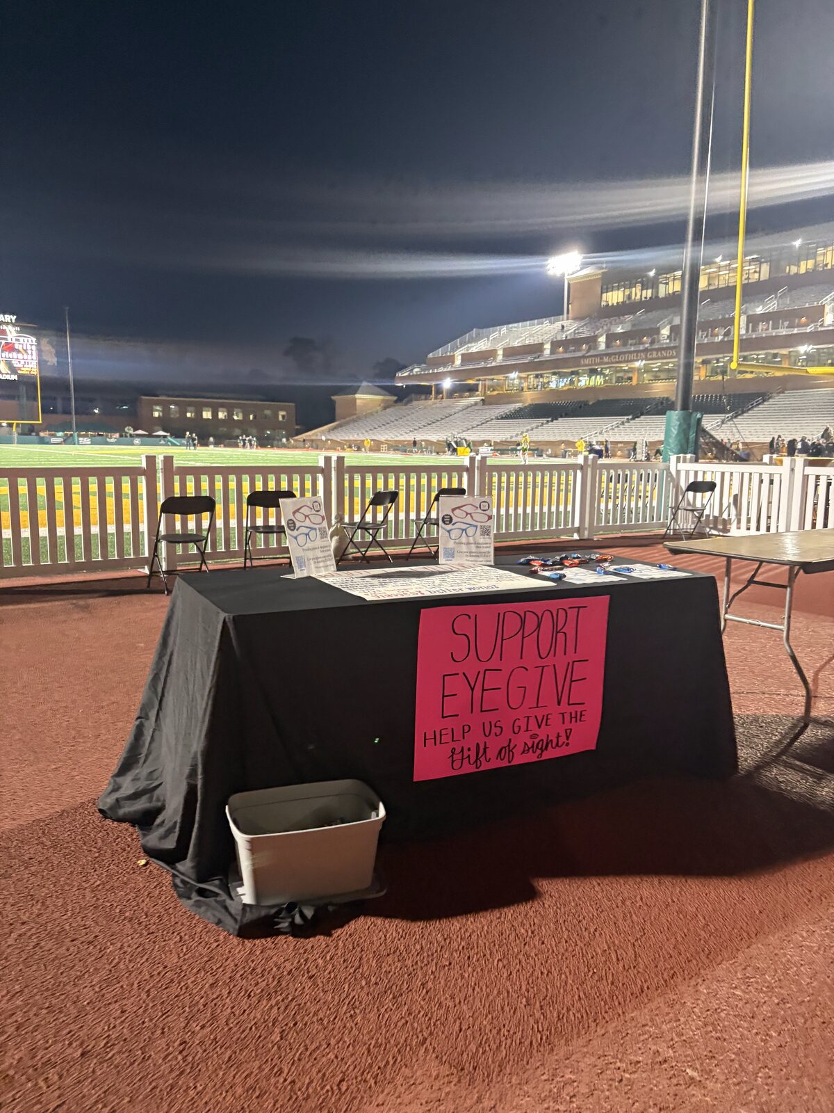 An EyeGive table set up beside the field at Zable Stadium during an evening William & Mary football game, with a pink &lsquo;Support EyeGive&rsquo; sign